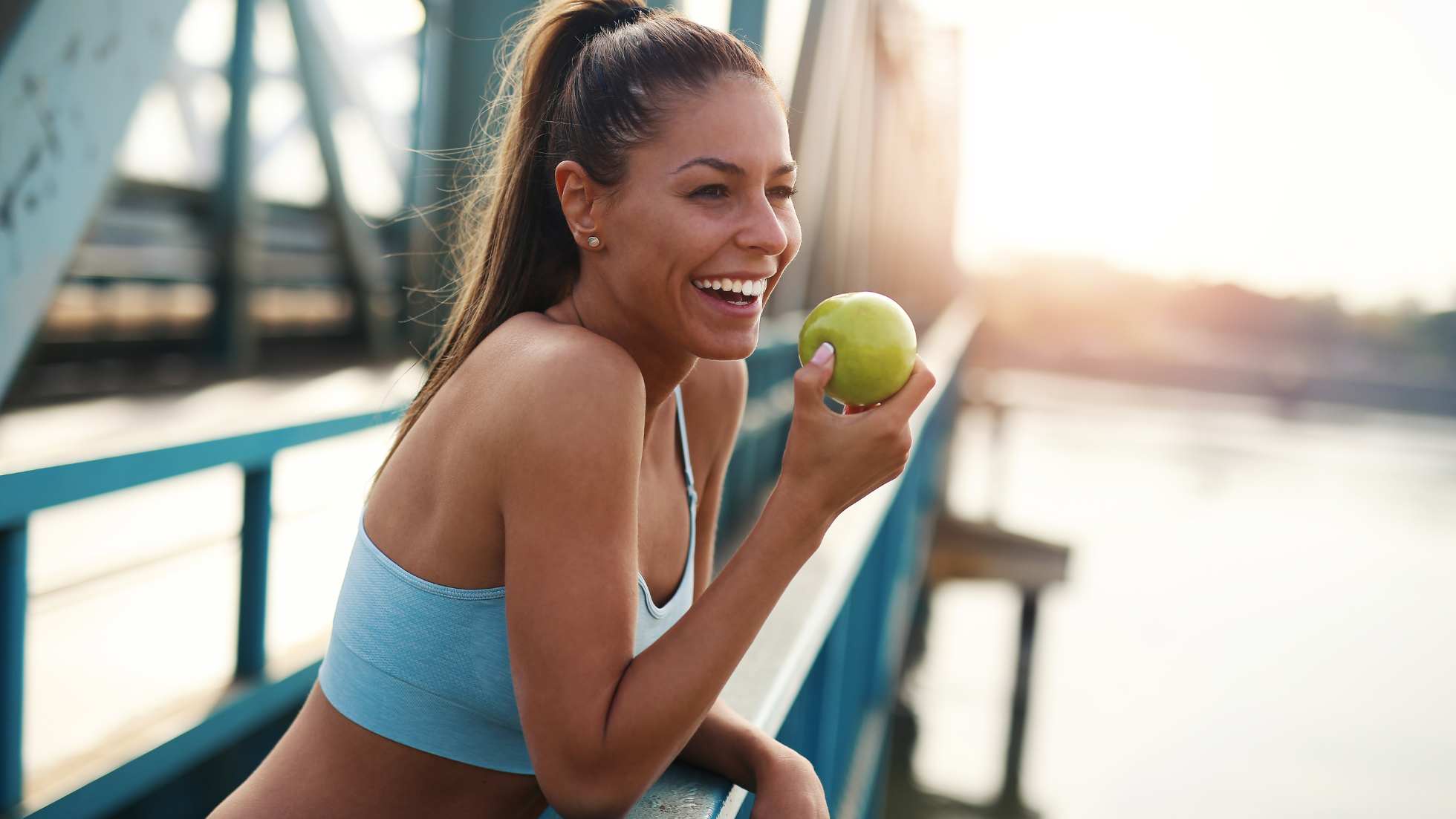 A joyful woman wearing a light blue sports bra is seated on a bridge railing, holding a green apple. With her hair tied back and a radiant smile, she represents a vibrant image of health and wellness. 